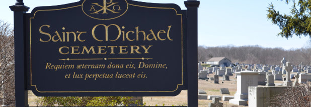 Large black Saint Michael Cemetery sign with gold letters with cemetery headstones in background