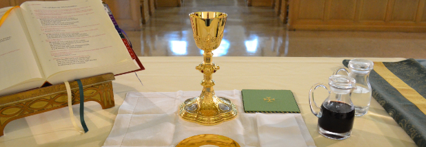 Large liturgy book, gold chalice, gold paten with communion host, and two cruets with wine and water on a white cloth on an altar with cream colored altar cloth, with light wooden church pews in background