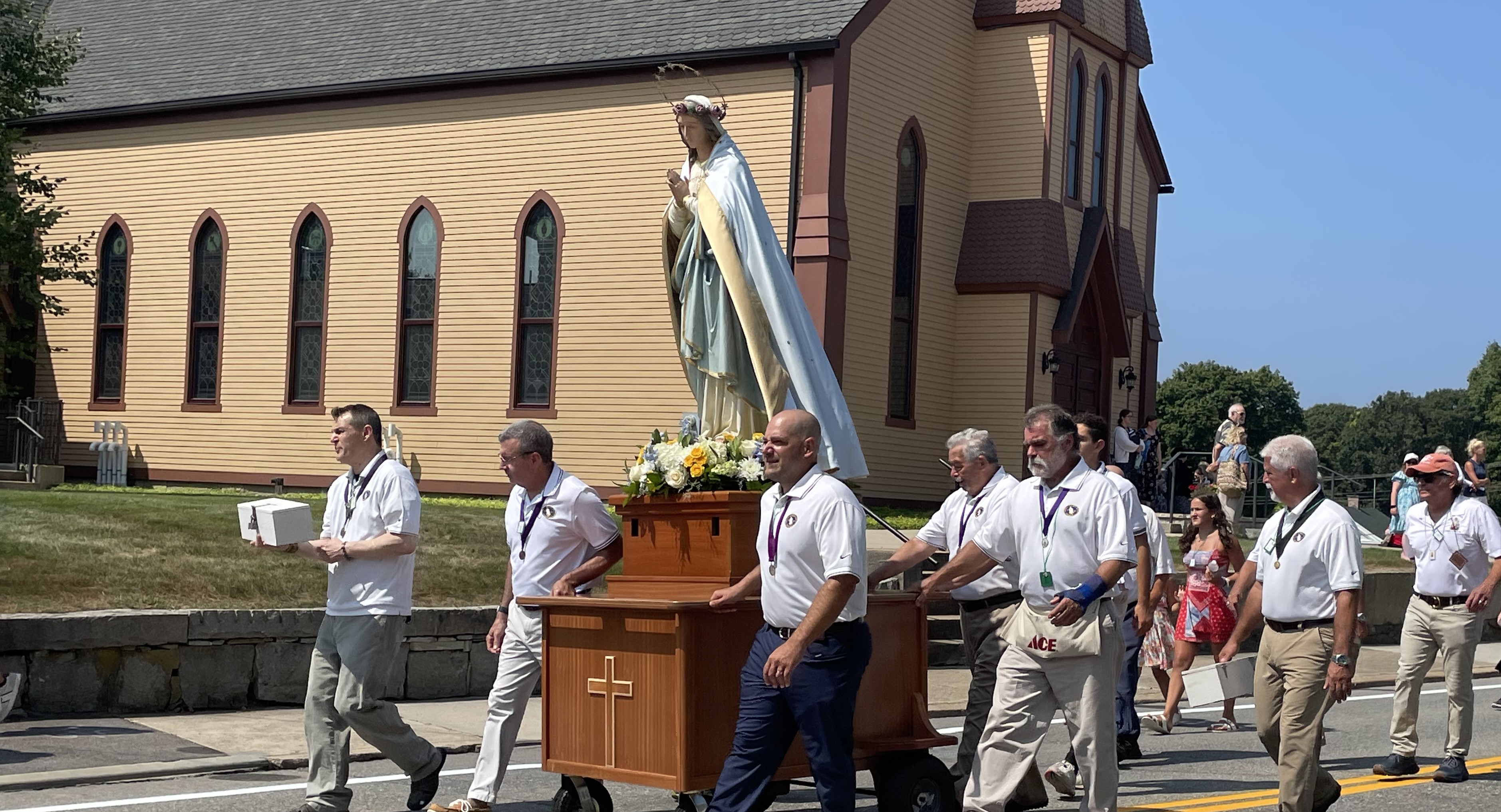 Statute of Blessed Mother with a crown of gold stars and light blue cape in parade with men in white shirts with large tan church in the background