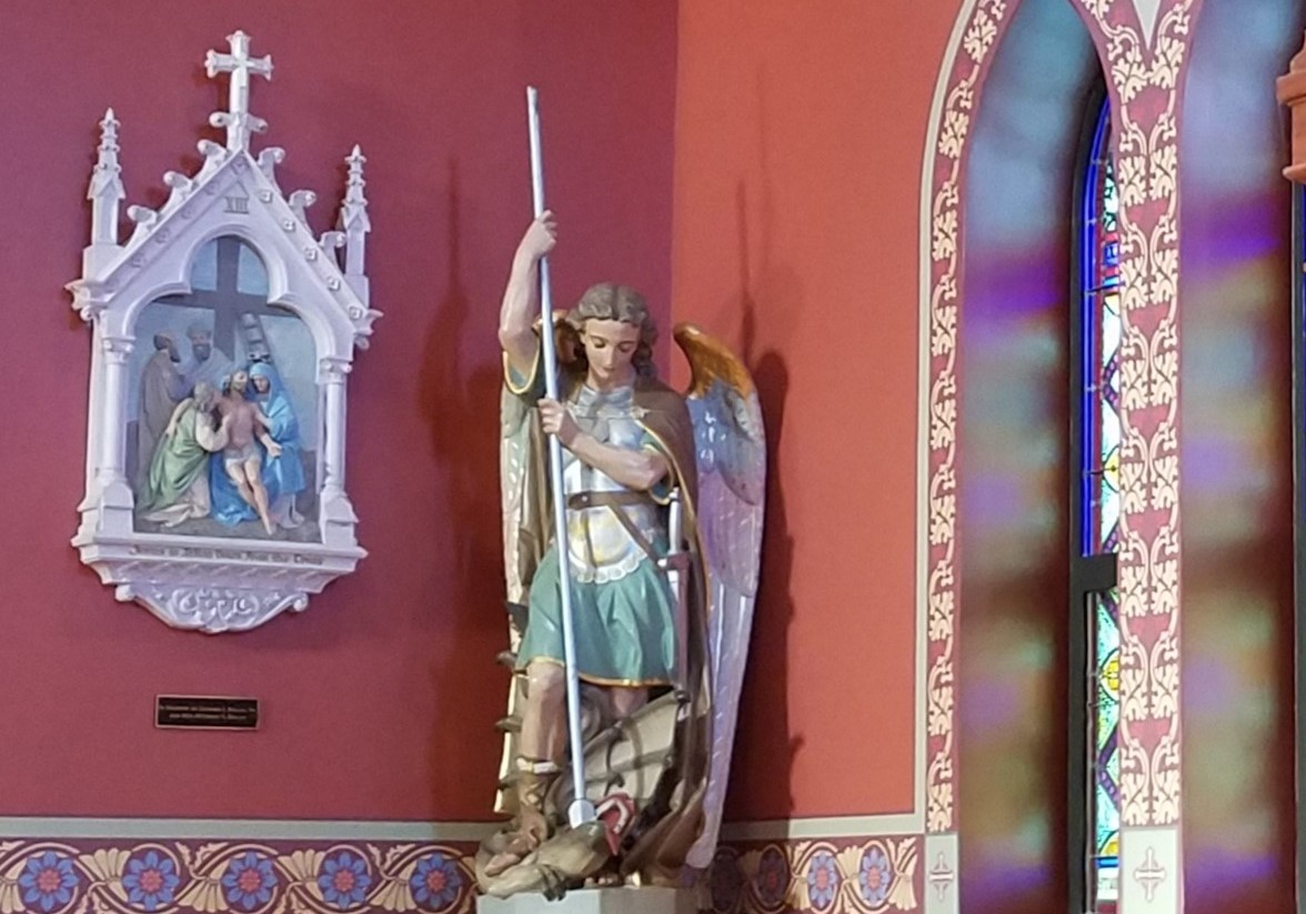 statue of St. Michael the Archangel in a church with a rust colored wall and station of the cross