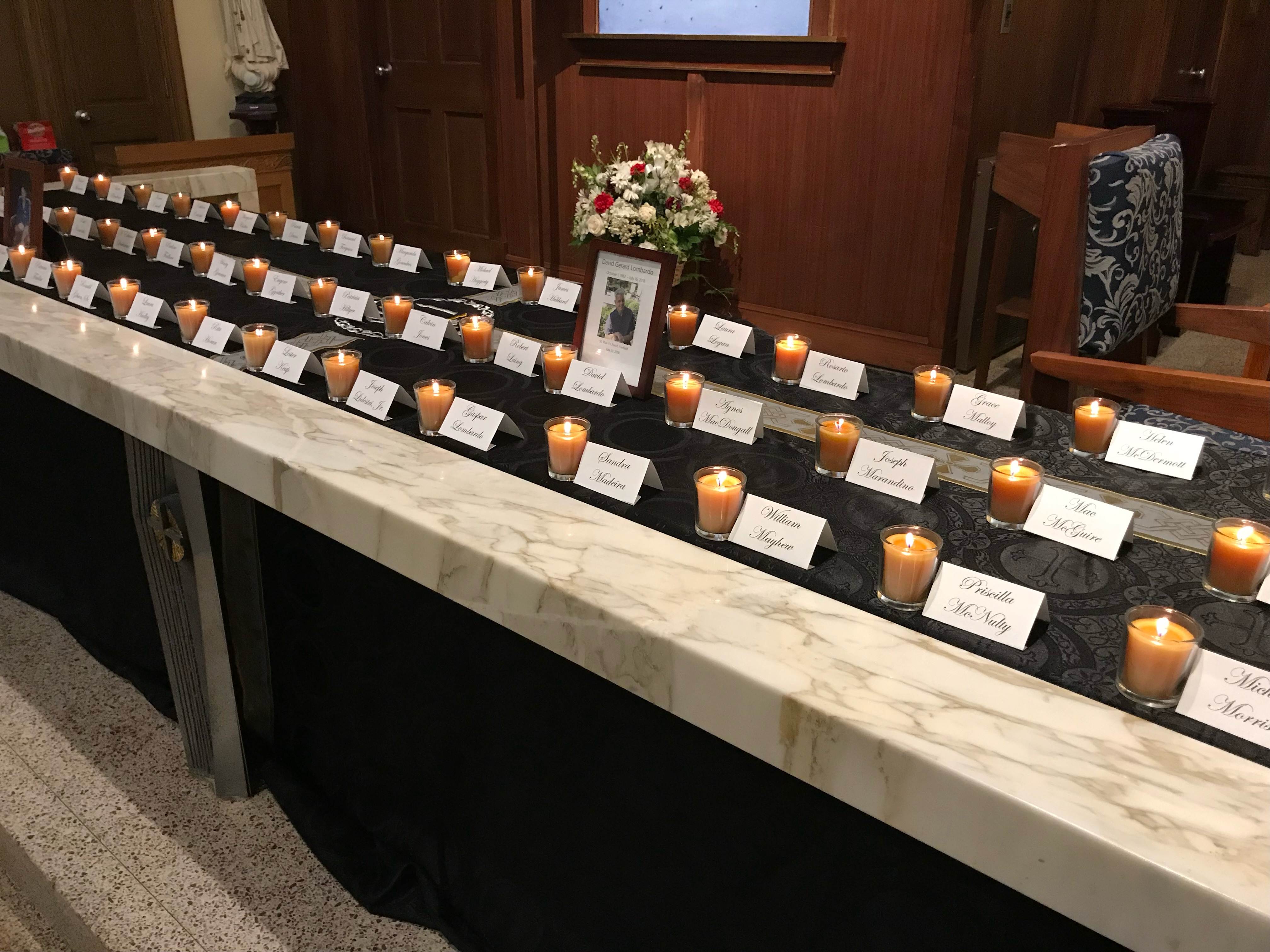 Photo of small lit candles next to name cards on long table with black table cloth behind marble rail